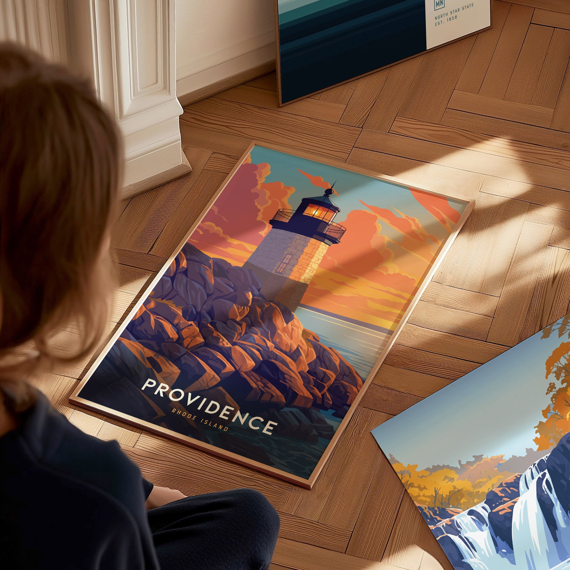 A person looks at a colorful poster of a lighthouse at sunset, labeled Providence Rhode Island, placed on a wooden floor. Another artwork is partially visible nearby.
