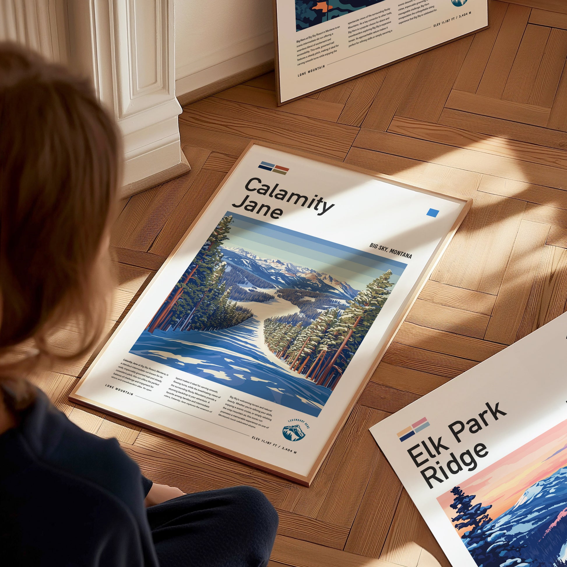 A person sits on a wooden floor, looking at a framed Calamity Jane ski run poster depicting a snowy path through pine trees with mountains in the background.