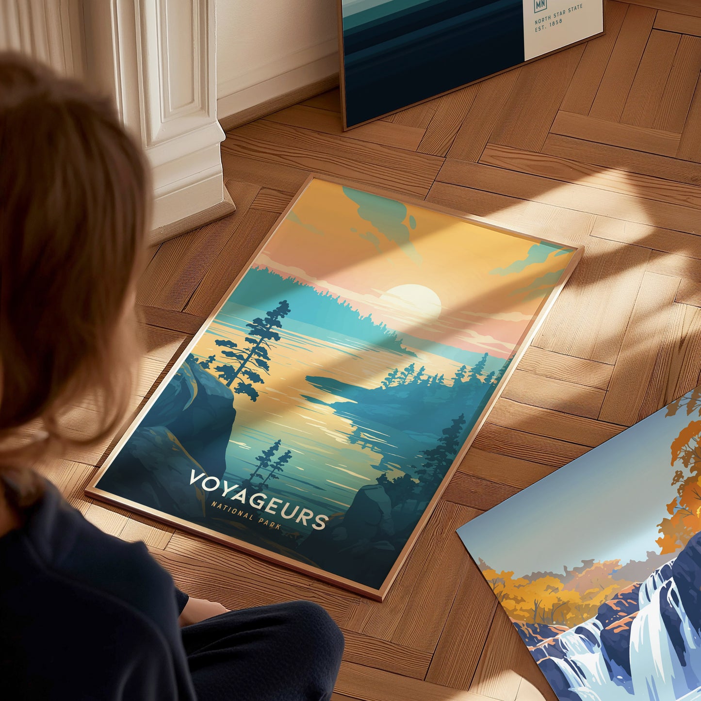 A person observes a vibrant poster of Voyageurs National Park on a wooden floor, showcasing a scenic sunset over a lake with silhouetted trees and rocks.