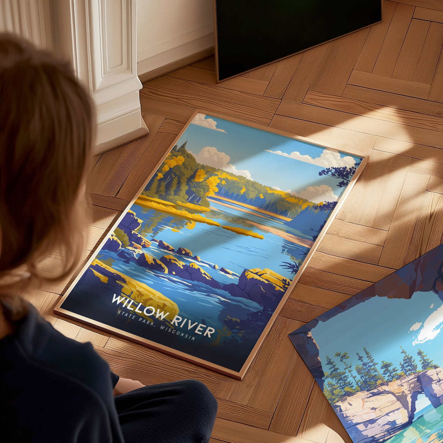 A child sits on a wooden floor, gazing at a vibrant travel print featuring Willow River State Park in Wisconsin.