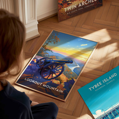 A person sits on a wooden floor, viewing a vibrant poster of Lookout Mountain featuring a cannon and scenic sunset. Nearby are posters of Twin Arches and Tybee Island.