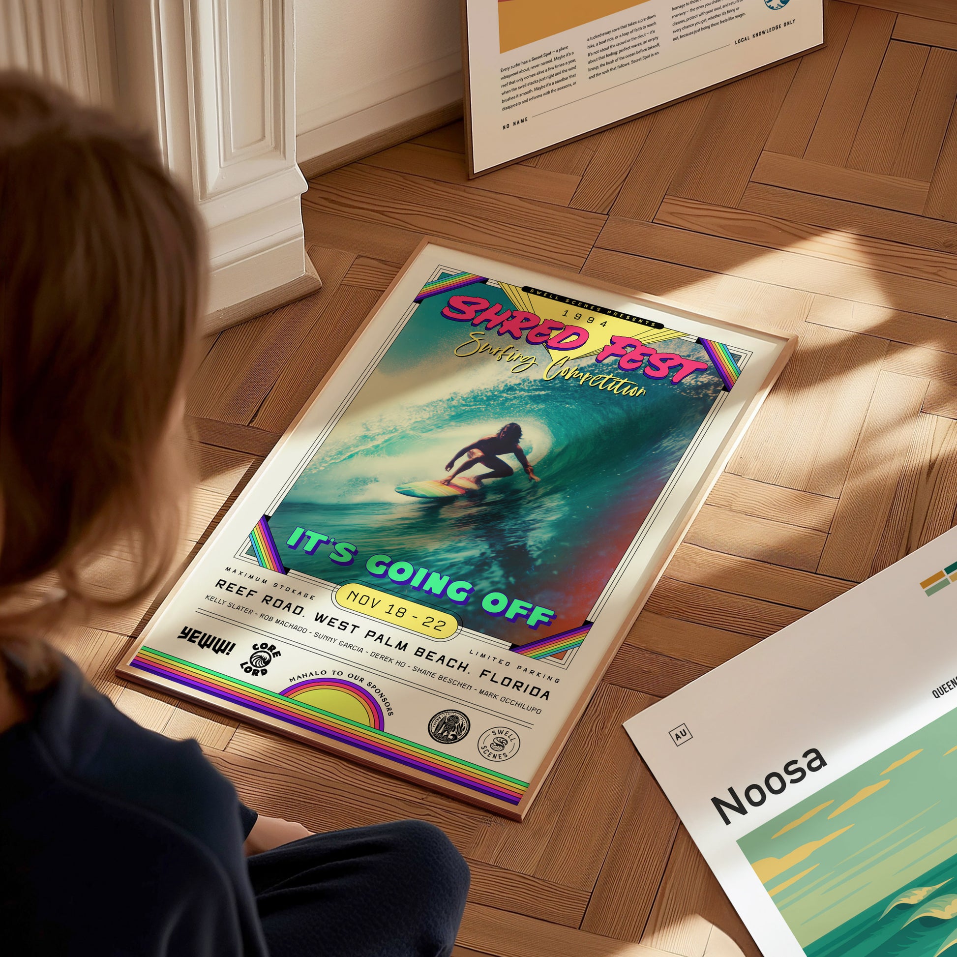 A child sits on a wooden floor, gazing at a colorful poster for SHRED FEST featuring a surfer riding a wave. The poster includes event details and vibrant graphics, surrounded by other framed artworks and posters in the background.