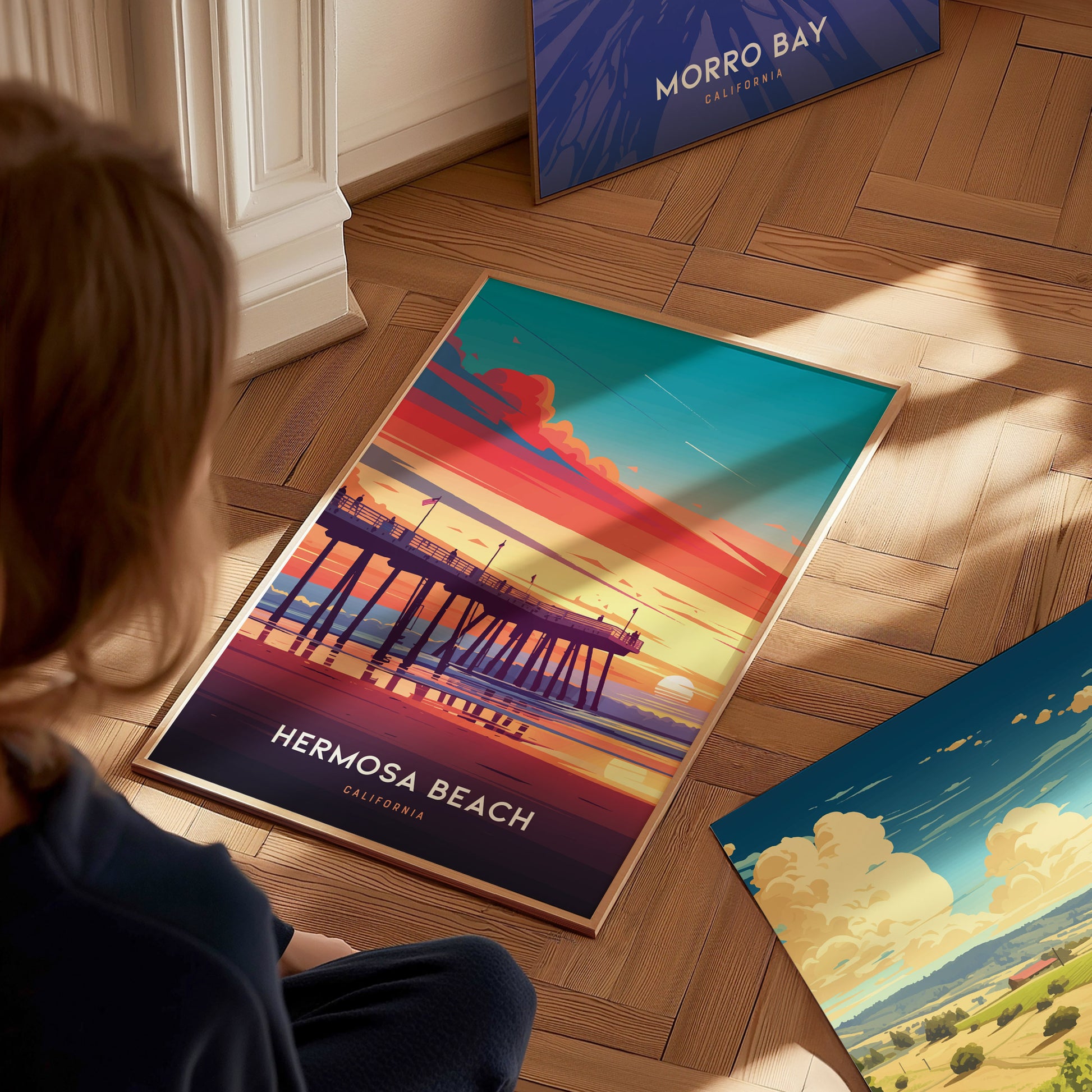 A person observes a colorful Hermosa Beach poster depicting a pier at sunset, with vibrant hues of orange, pink, and blue. Nearby, a Morro Bay poster is partially visible.