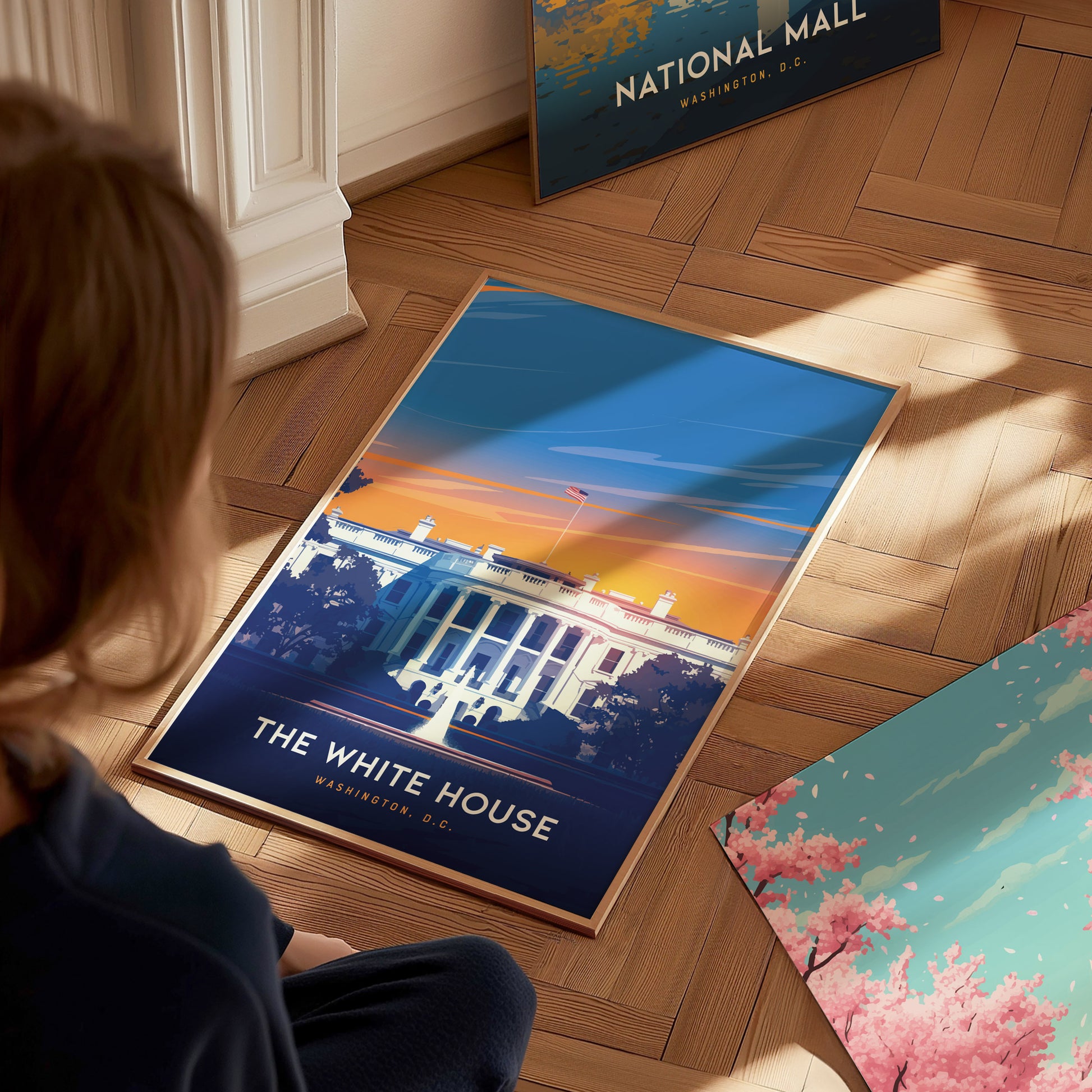 A person is looking at a White House poster on a wooden floor, featuring vibrant colors and bold lines. Nearby, another poster partially shows cherry blossoms.