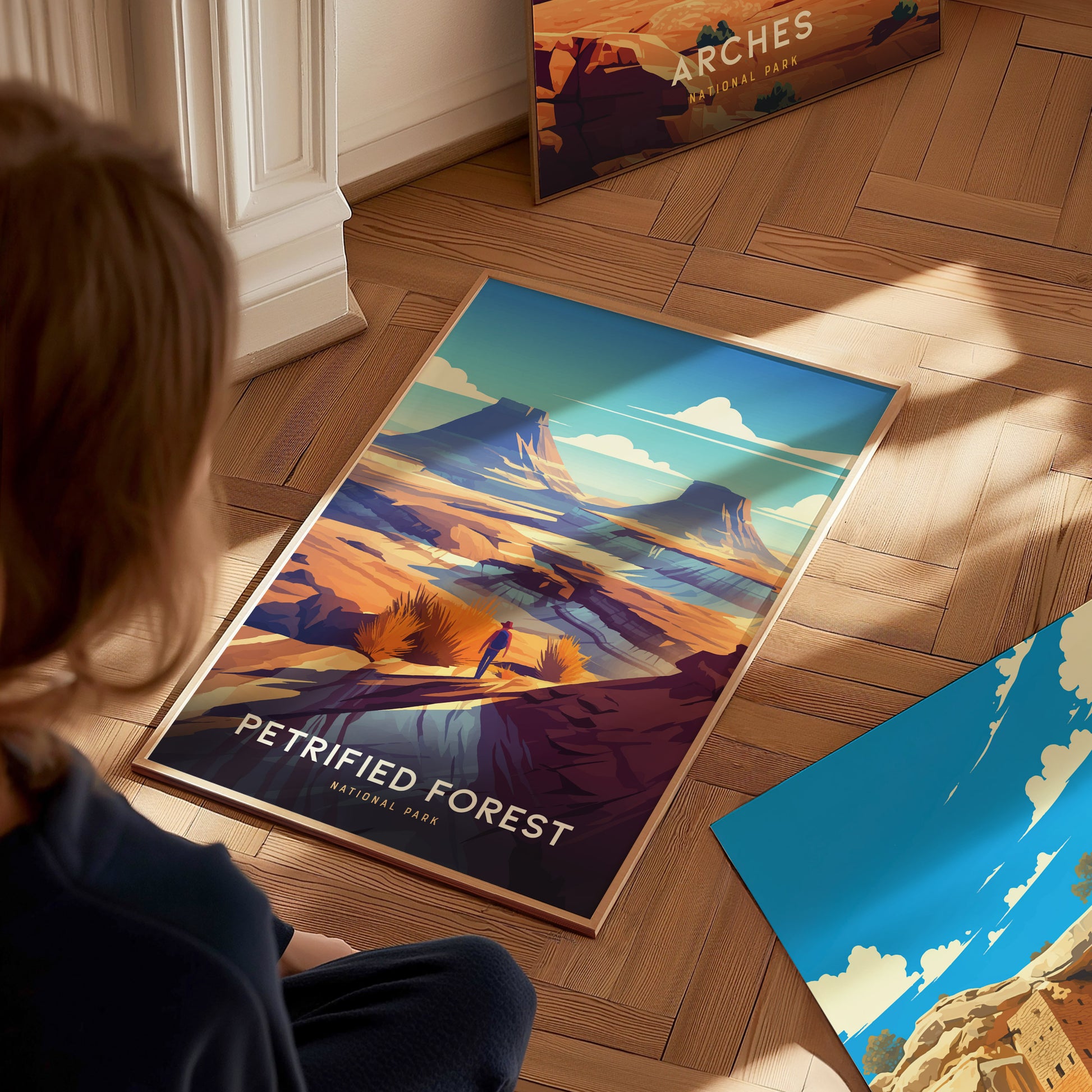 A person sits on a wooden floor, admiring a colorful Petrified Forest National Park poster depicting a desert landscape with mountains and clouds. Another poster is partially visible nearby.