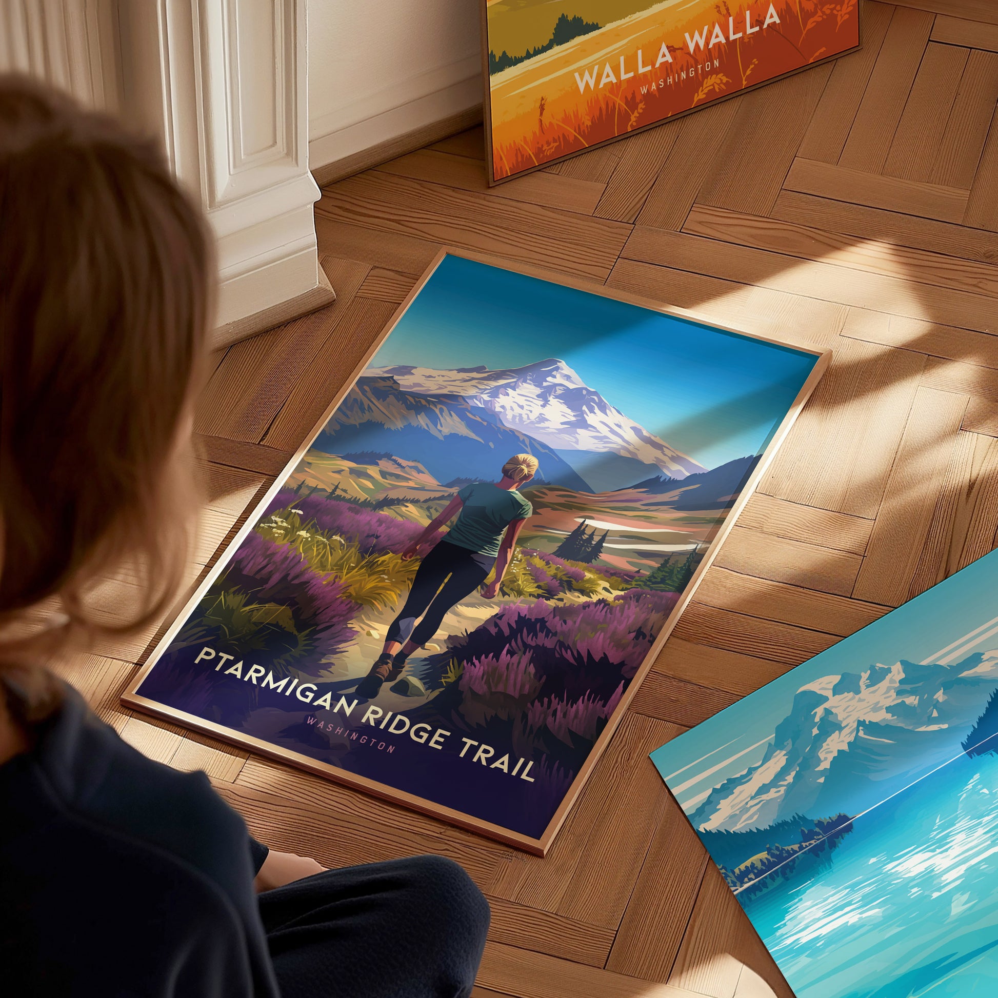 A person observes a vibrant poster of Ptarmigan Ridge Trail featuring a hiker amidst wildflowers with Mt. Baker in the background, placed on a wooden floor alongside other artwork.