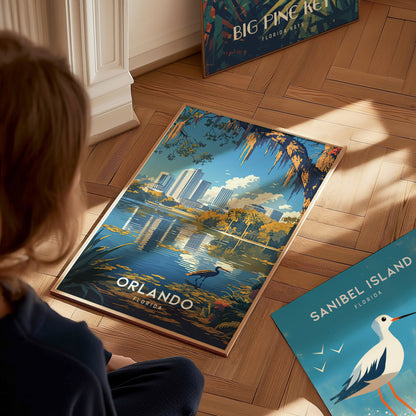 A person observes a vibrant poster of Orlando, Florida, featuring a lake with a heron, city skyline, and lush foliage, placed on a wooden floor. Nearby, another poster is visible.