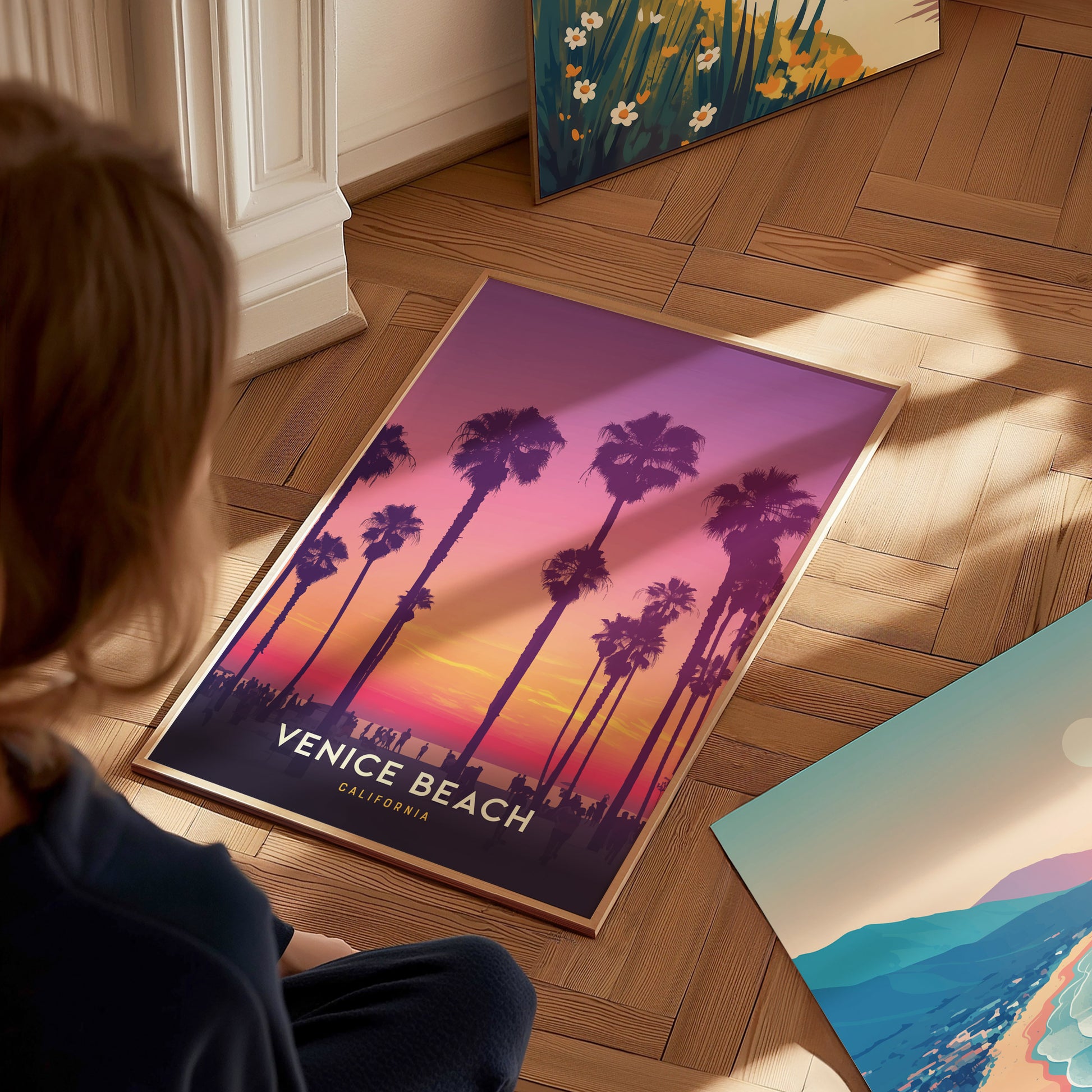 A person observes a framed Venice Beach poster on a wooden floor, featuring palm trees silhouetted against a vibrant sunset in shades of purple, pink, and orange.