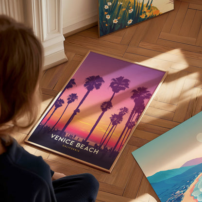 A person observes a framed Venice Beach poster on a wooden floor, featuring palm trees silhouetted against a vibrant sunset in shades of purple, pink, and orange.