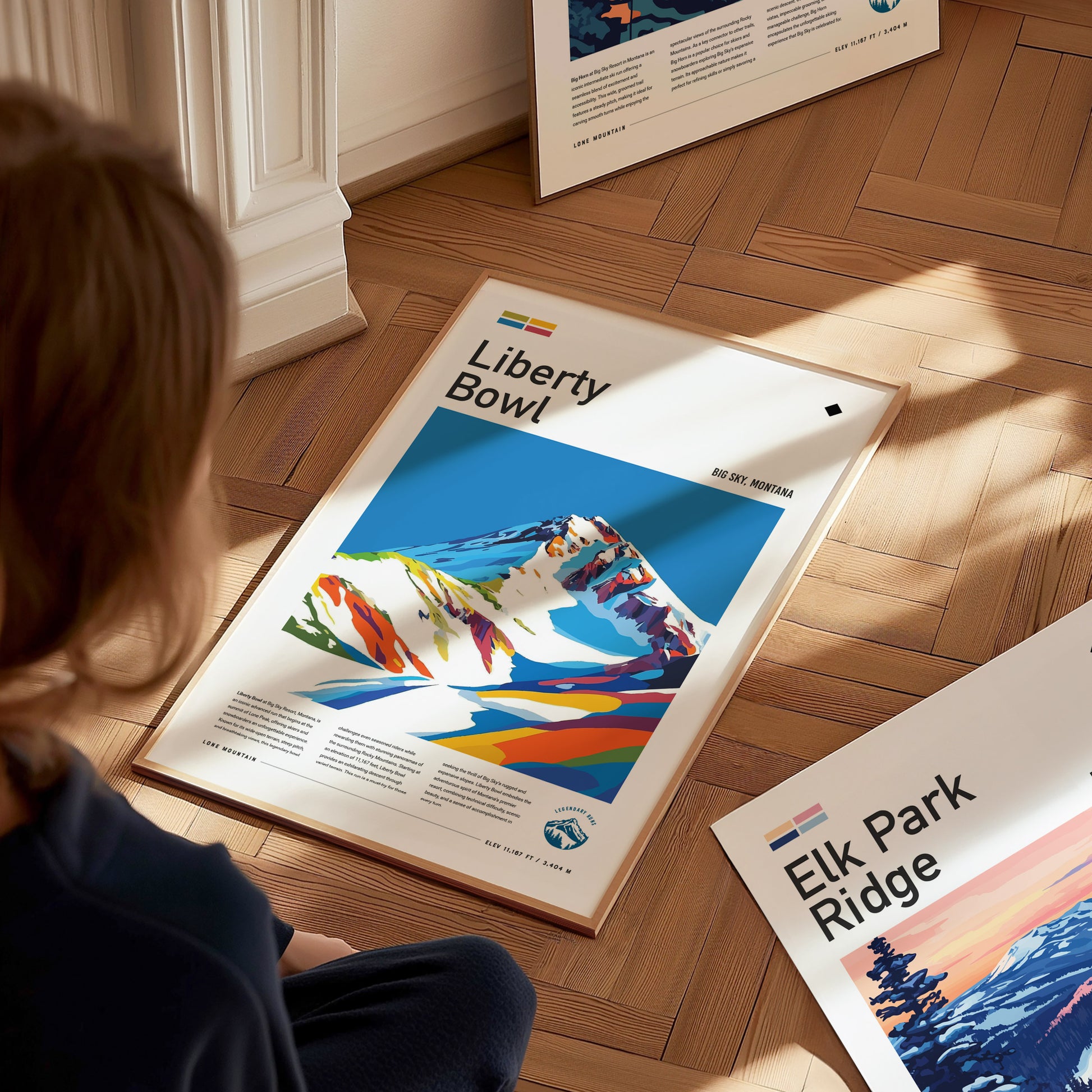 A person looks at a vibrant Liberty Bowl poster featuring colorful, abstract mountain art. The poster lies on a wooden floor next to an Elk Park Ridge print.