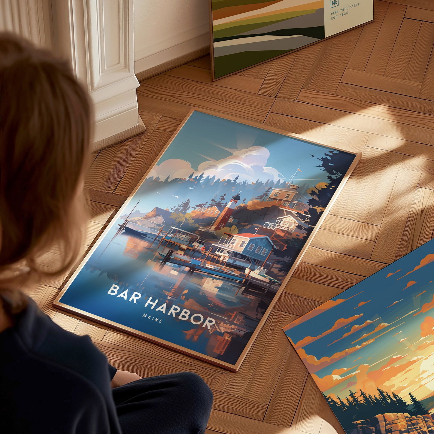 A person sits on a wooden floor, looking at a framed Bar Harbor, Maine poster depicting a coastal scene with houses and boats. Sunlight casts shadows on the floor.