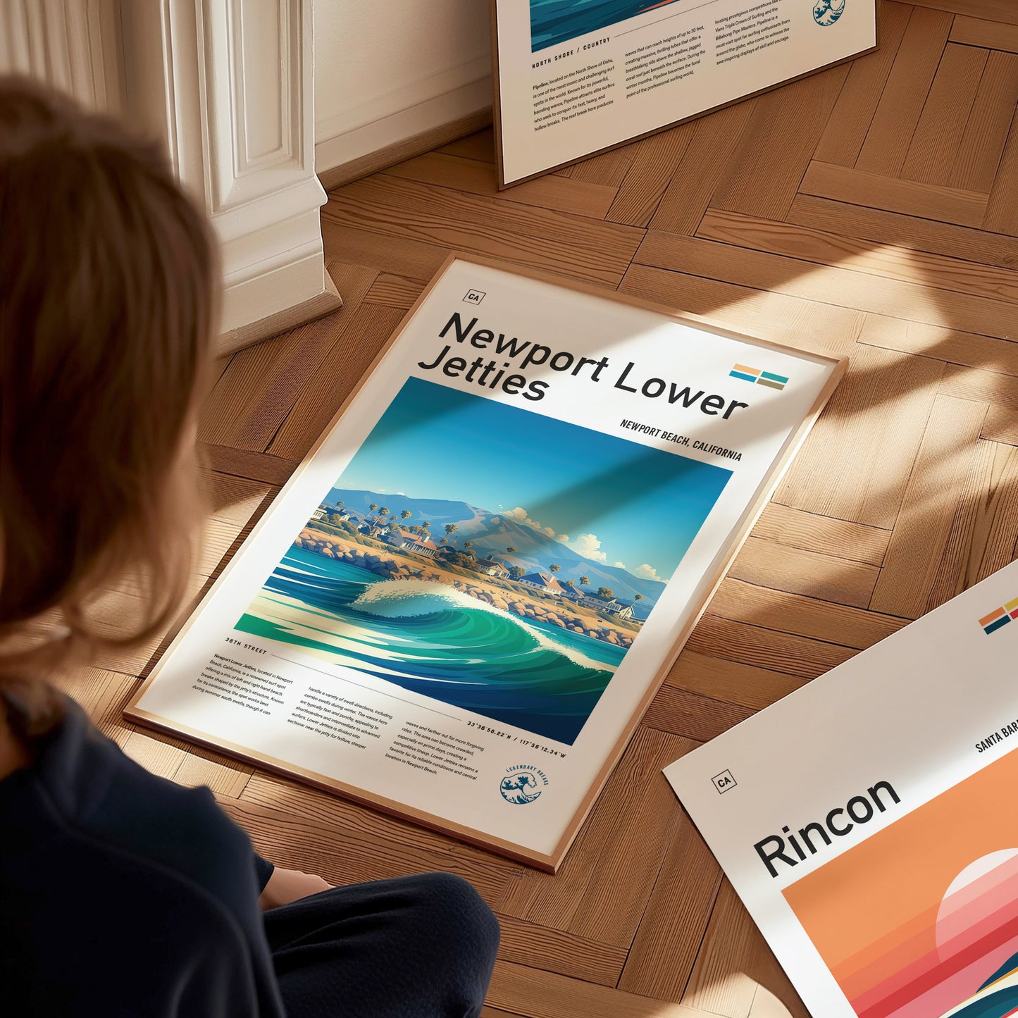 A person sits on a wooden floor, looking at a framed Newport Lower Jetties poster depicting a vibrant beach scene. Nearby, another framed poster labeled Rincon is partially visible.