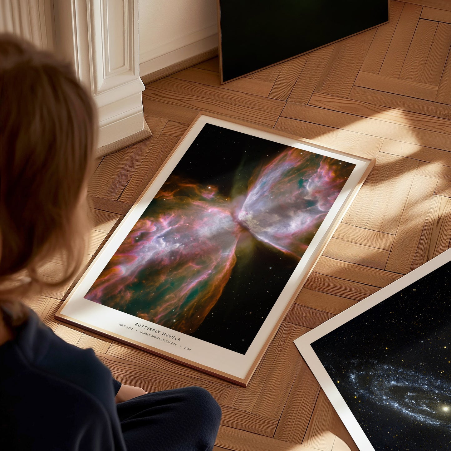 A child sits on a hardwood floor, gazing at a vibrant poster of the Butterfly Nebula, showcasing colorful cosmic clouds and star formations. Nearby, another poster of a galaxy is partially visible, enhancing the celestial theme.