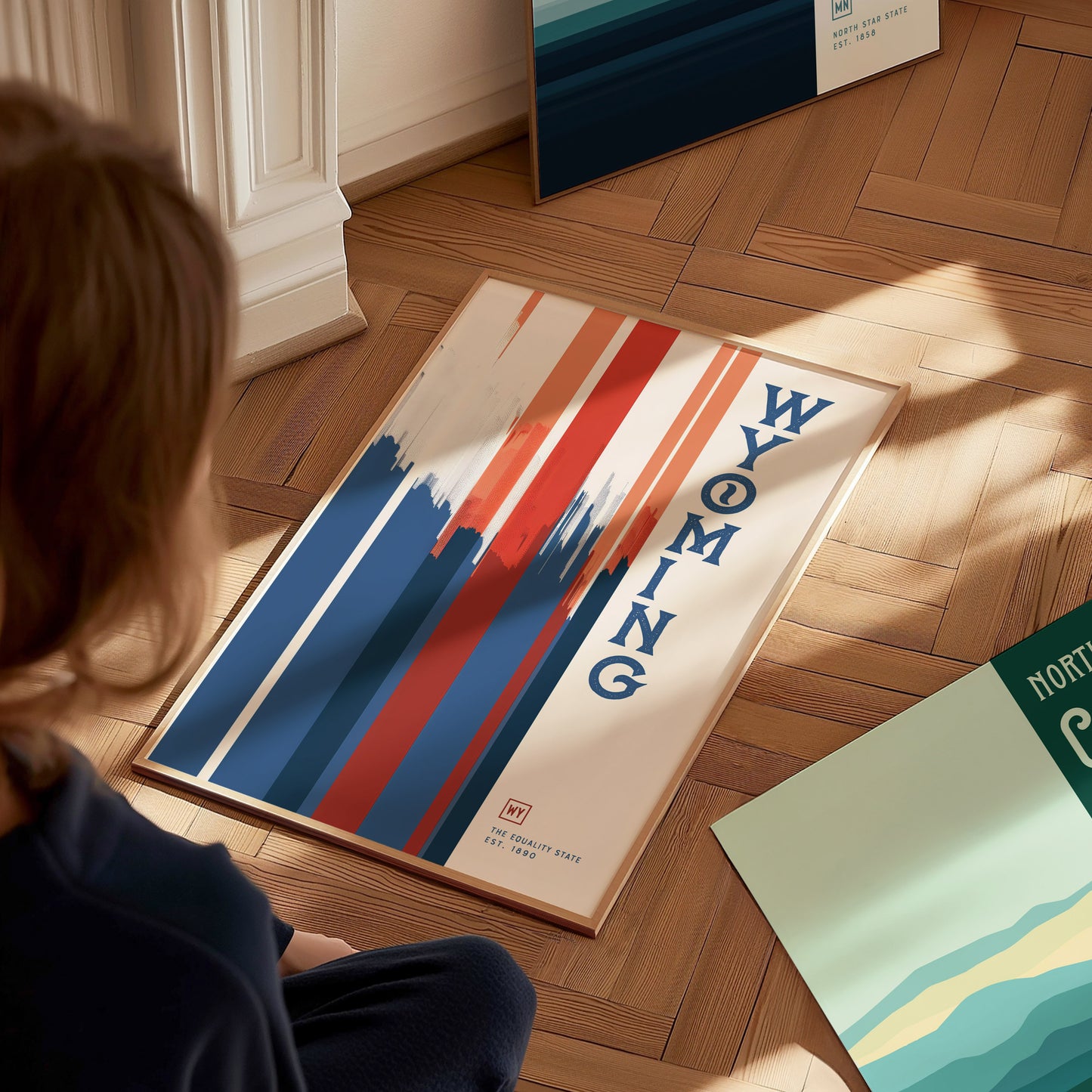 A person sits on a wooden floor, looking at a framed Wyoming poster featuring bold red, white, and blue vertical stripes with the word Wyoming written vertically.