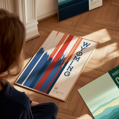 A person sits on a wooden floor, looking at a framed Wyoming poster featuring bold red, white, and blue vertical stripes with the word Wyoming written vertically.