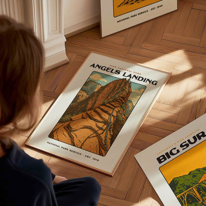 A person sits on a wooden floor gazing at a vintage Angels Landing poster depicting a steep sandstone trail. Another poster titled Big Sur is partially visible nearby.
