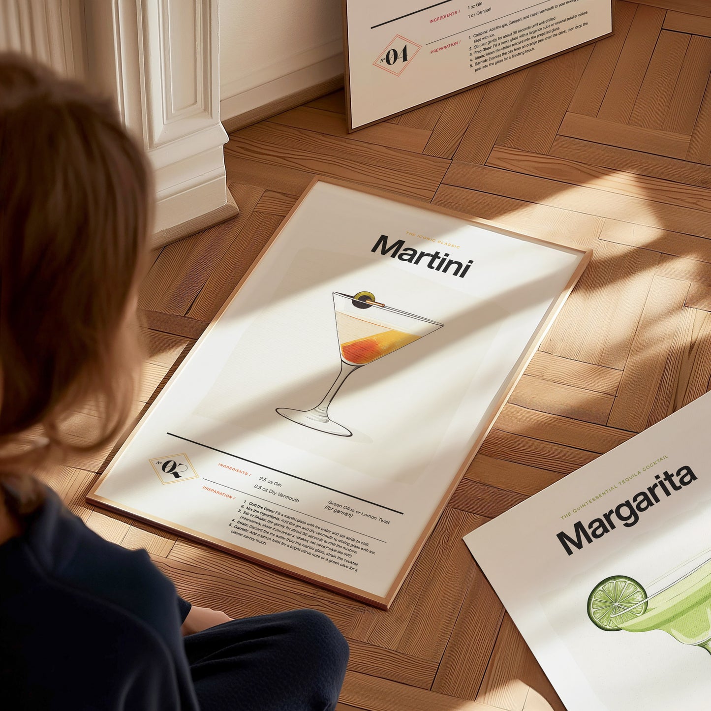A person sits on a wooden floor, looking at a Martini cocktail poster displaying a glass with an olive. Nearby, a Margarita poster is partially visible.