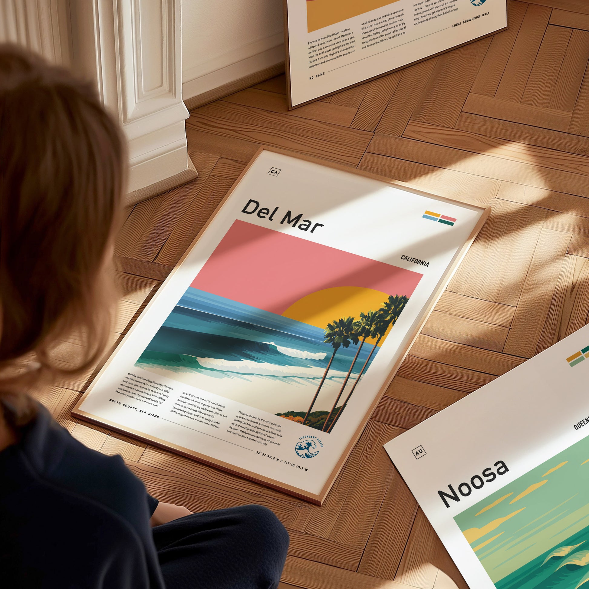 A child sits on a wooden floor, gazing at a framed poster titled Del Mar, depicting a vibrant sunset over a beach scene with palm trees and gentle waves. Other posters titled Noosa and other designs are visible in the background.