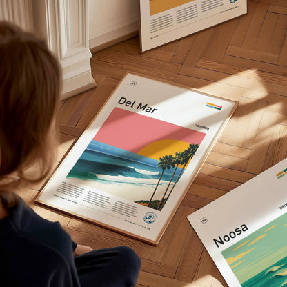 A child sits on a wooden floor, gazing at a framed poster titled Del Mar, depicting a vibrant sunset over a beach scene with palm trees and gentle waves. Other posters titled Noosa and other designs are visible in the background.