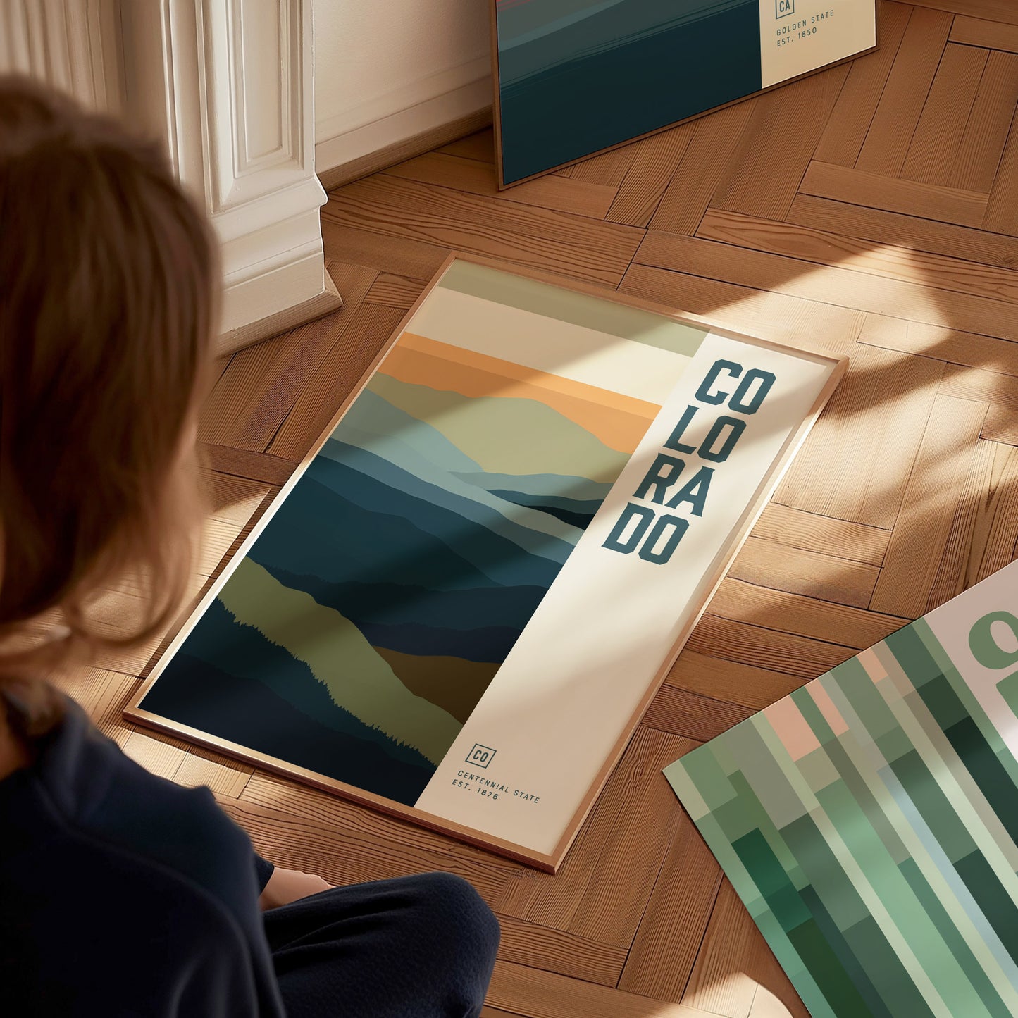 A person sits on a wooden floor, looking at a framed poster depicting abstract layers of blue and green mountains with the text COLORADO vertically aligned. Sunlight casts soft shadows.