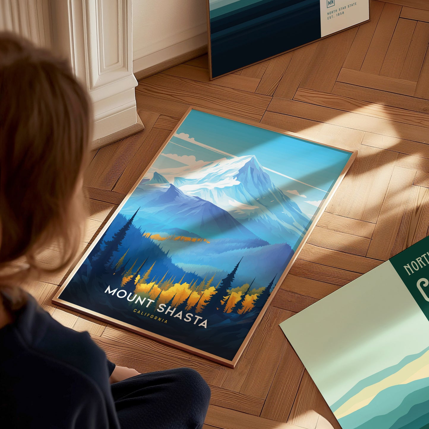A person views a vibrant Mount Shasta poster on a wooden floor, featuring a snow-capped mountain, forested foothills, and a clear sky. Additional posters are partially visible nearby.