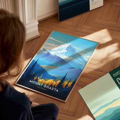A person views a vibrant Mount Shasta poster on a wooden floor, featuring a snow-capped mountain, forested foothills, and a clear sky. Additional posters are partially visible nearby.