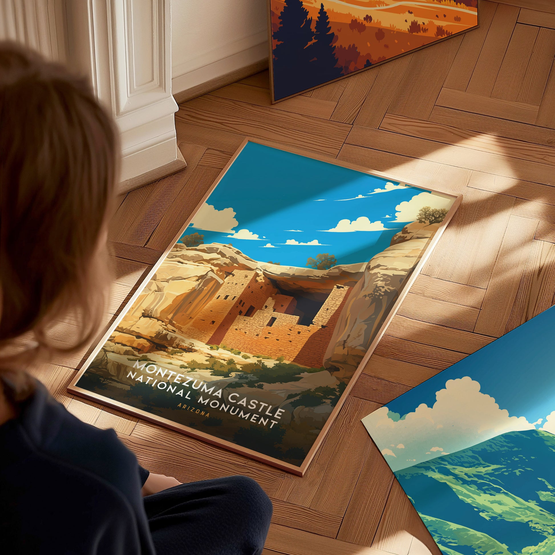 A person sits on a wooden floor, looking at a vibrant Montezuma Castle National Monument poster featuring cliff dwellings under a bright blue sky. Other posters are partially visible nearby.