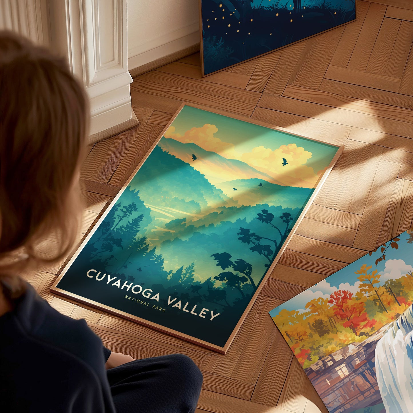 A person views a Cuyahoga Valley National Park poster on a wooden floor, featuring vibrant blue and green hills, trees, and birds against a sunset sky.