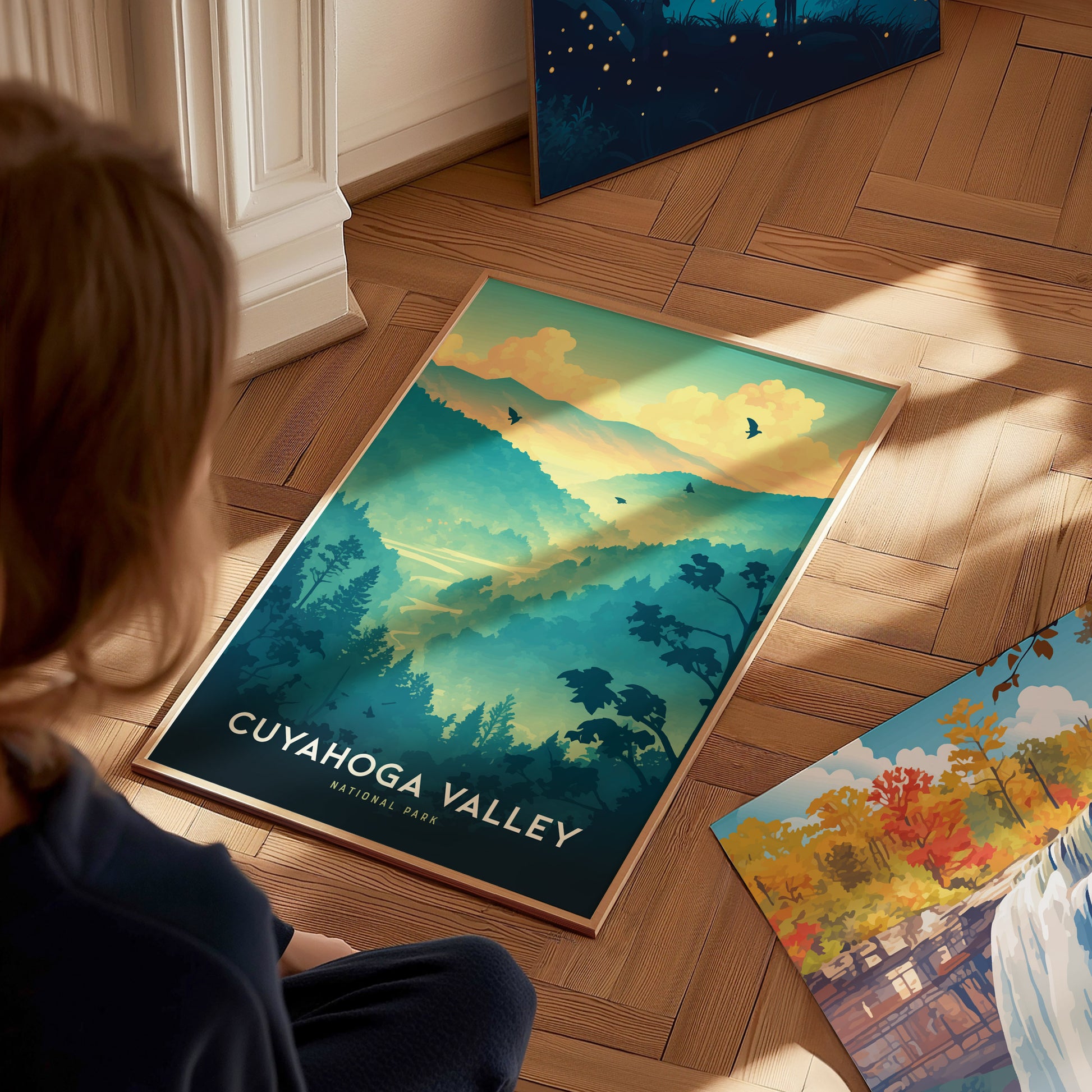 A person views a Cuyahoga Valley National Park poster on a wooden floor, featuring vibrant blue and green hills, trees, and birds against a sunset sky.
