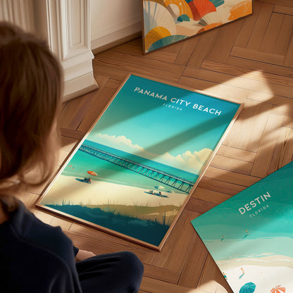 A person is sitting on a wooden floor, looking at a framed Panama City Beach poster featuring a beach, ocean, and pier. Another Destin, Florida poster is nearby.