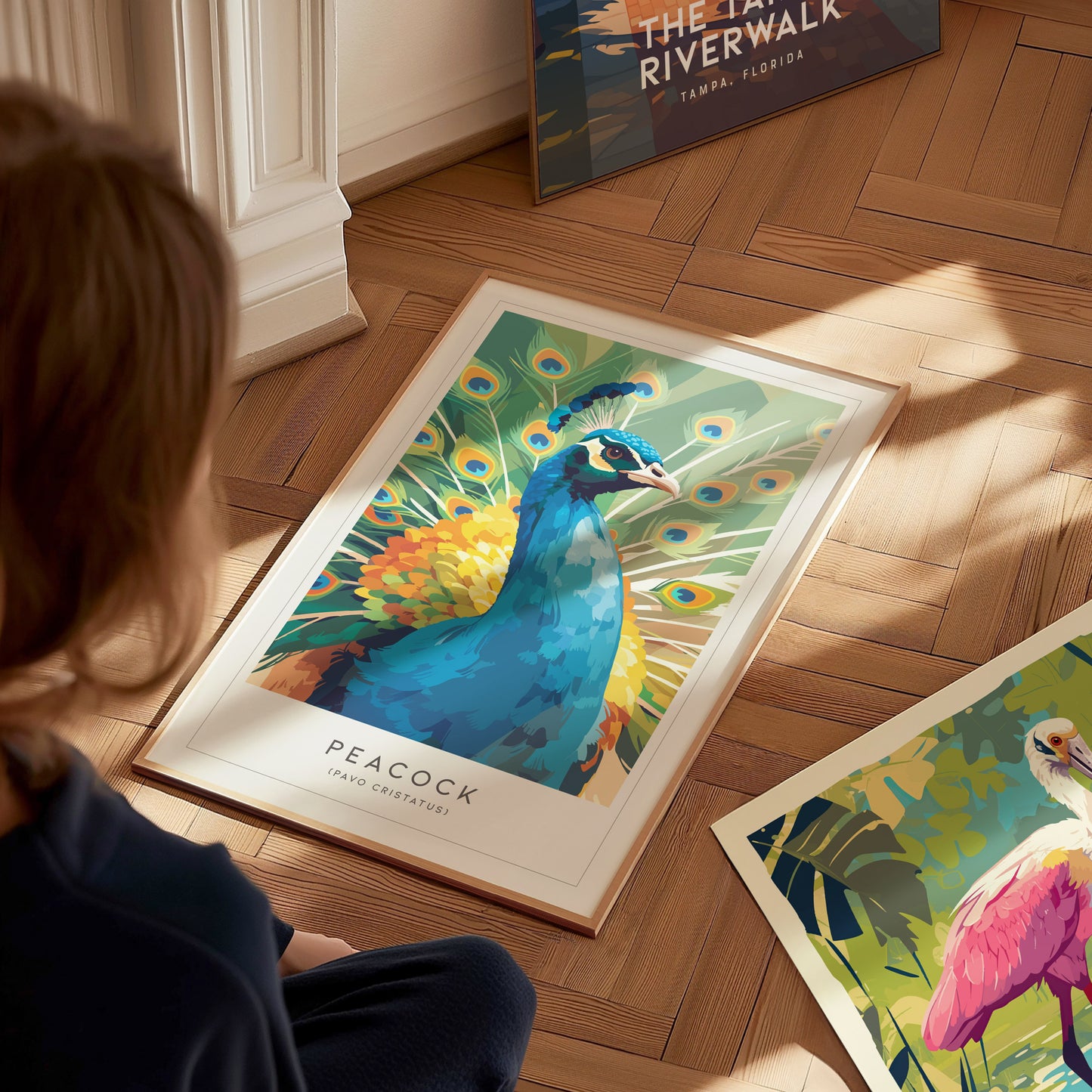 A vibrant, colorful poster of a peacock displays its striking plumage, featuring intricate patterns and bright hues. The artwork is placed on a wooden floor, surrounded by additional posters, creating a lively and artistic atmosphere.