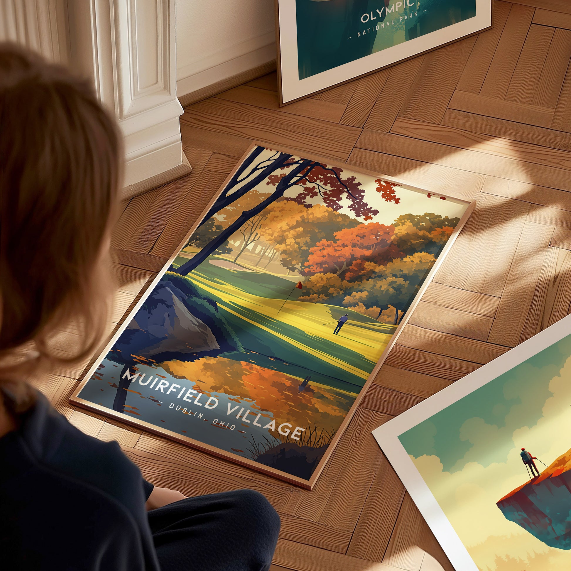 A person looks at a vividly colored Muirfield Village Golf Club poster on a wooden floor, featuring autumn trees and a golfer on the course.