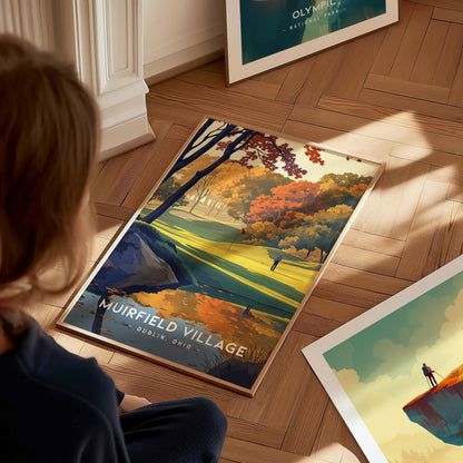 A person looks at a vividly colored Muirfield Village Golf Club poster on a wooden floor, featuring autumn trees and a golfer on the course.