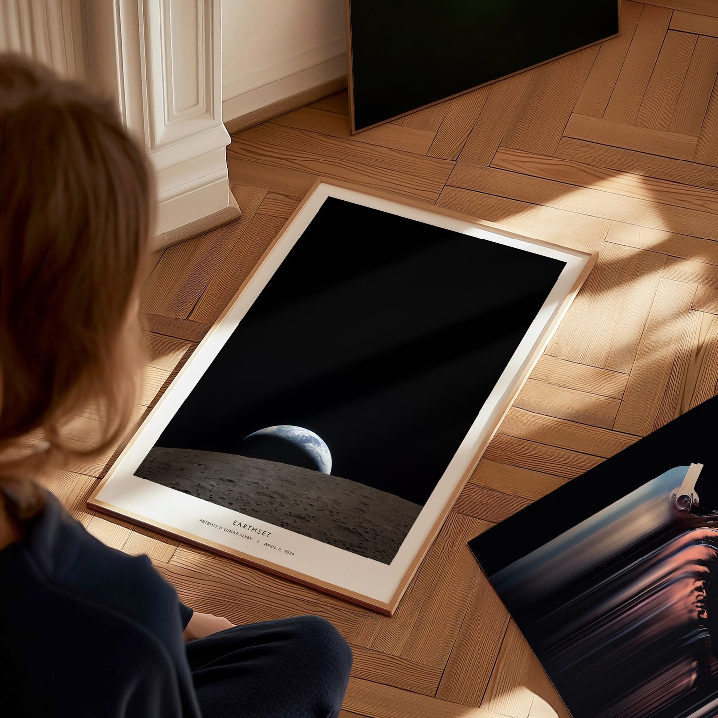 A child sits on a wooden floor, gazing at a poster featuring a view of Earth from the Moons surface, with a dark, space backdrop. The poster is framed and partially propped against a wall, surrounded by other artworks.