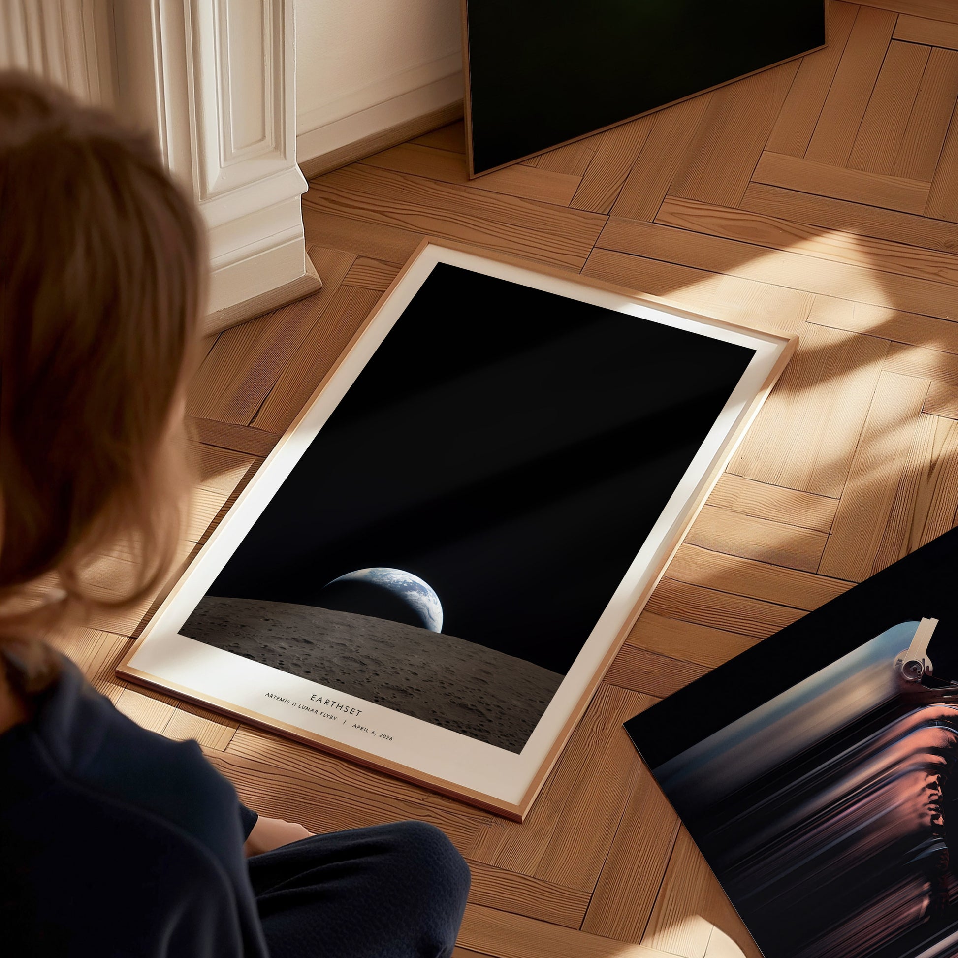 A child sits on a wooden floor, gazing at a poster featuring a view of Earth from the Moons surface, with a dark, space backdrop. The poster is framed and partially propped against a wall, surrounded by other artworks.