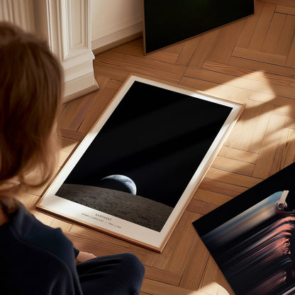 A child sits on a wooden floor, gazing at a poster featuring a view of Earth from the Moons surface, with a dark, space backdrop. The poster is framed and partially propped against a wall, surrounded by other artworks.