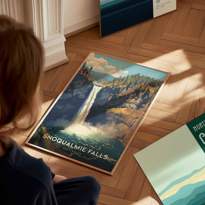 A person views a framed Snoqualmie Falls poster on a wooden floor, depicting a waterfall surrounded by lush greenery and dramatic landscapes. Nearby, partial views of other posters are visible.