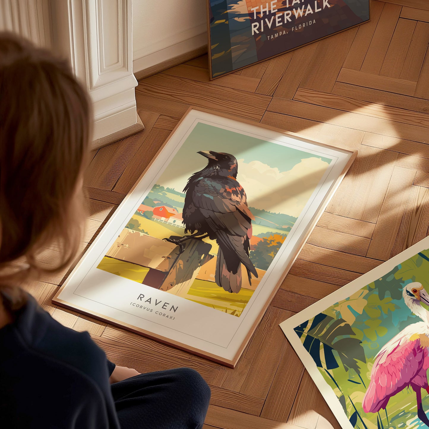 A child sits on a wooden floor, examining a framed print of a raven perched on a branch, with a colorful landscape in the background. Several other art prints, including one featuring a flamingo, are scattered nearby.