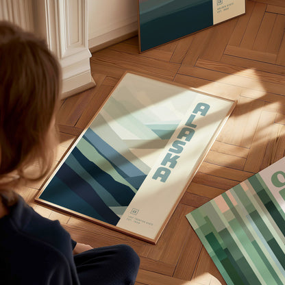 A person observes a minimalist poster featuring abstract mountains and the word Alaska on a wooden floor. Sunlight casts soft shadows on the artwork.