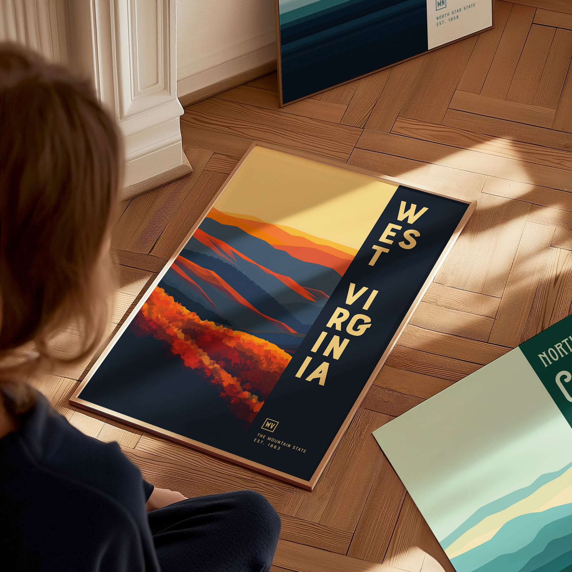 A person looks at a vibrant West Virginia poster on a wooden floor, featuring colorful abstract mountains and bold text. Other posters are partially visible nearby.