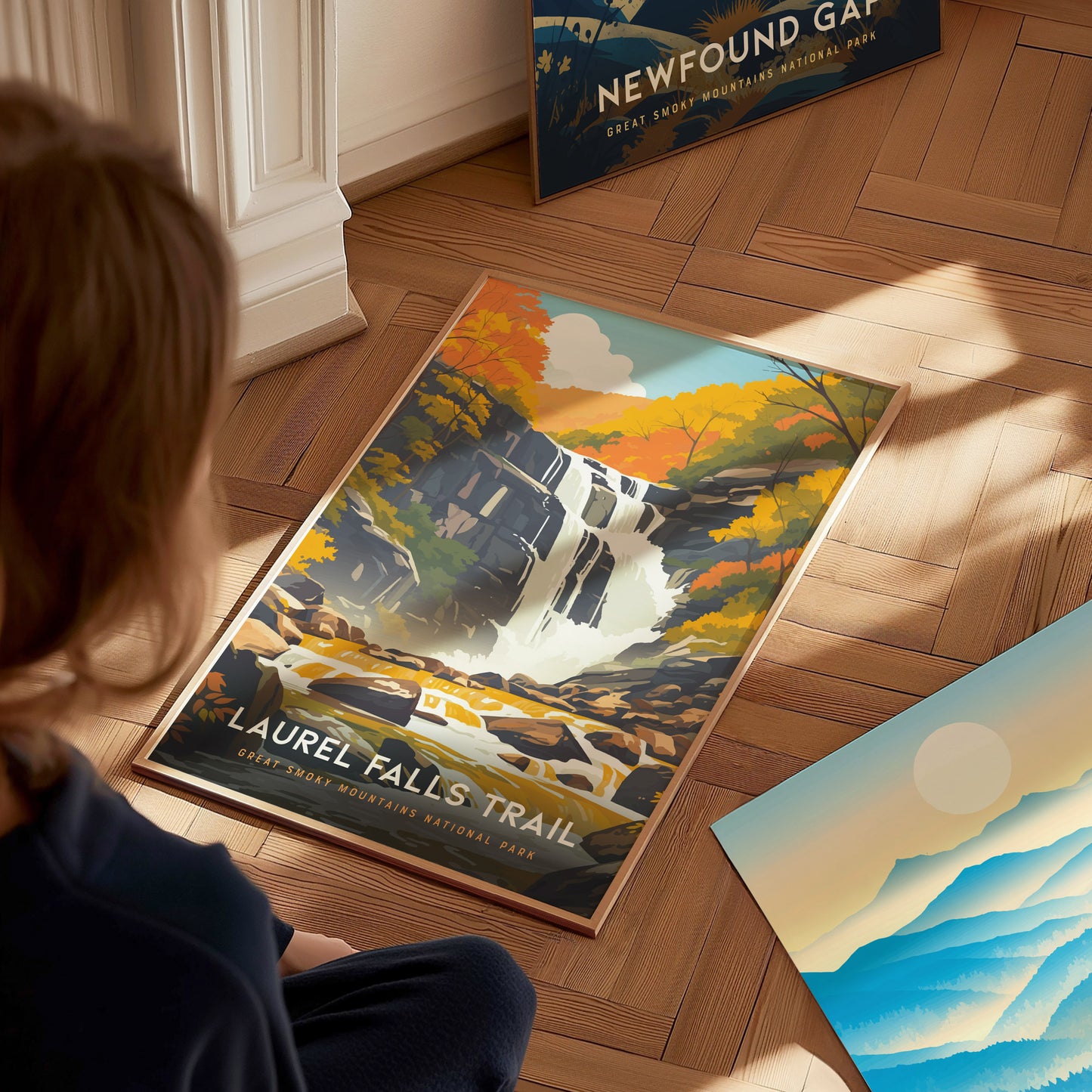 A person observes a colorful poster of Laurel Falls Trail featuring a waterfall amidst autumn foliage, placed on a wooden floor. Other posters are partially visible nearby.