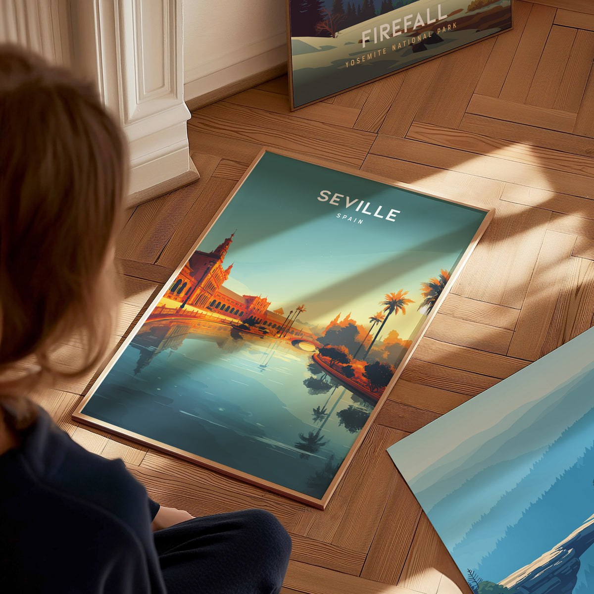 A child sits on a wooden floor, gazing at a vibrant poster featuring a stylized view of Seville, Spain. The artwork showcases golden towers reflecting in calm water, framed by palm trees and a serene sunset sky.