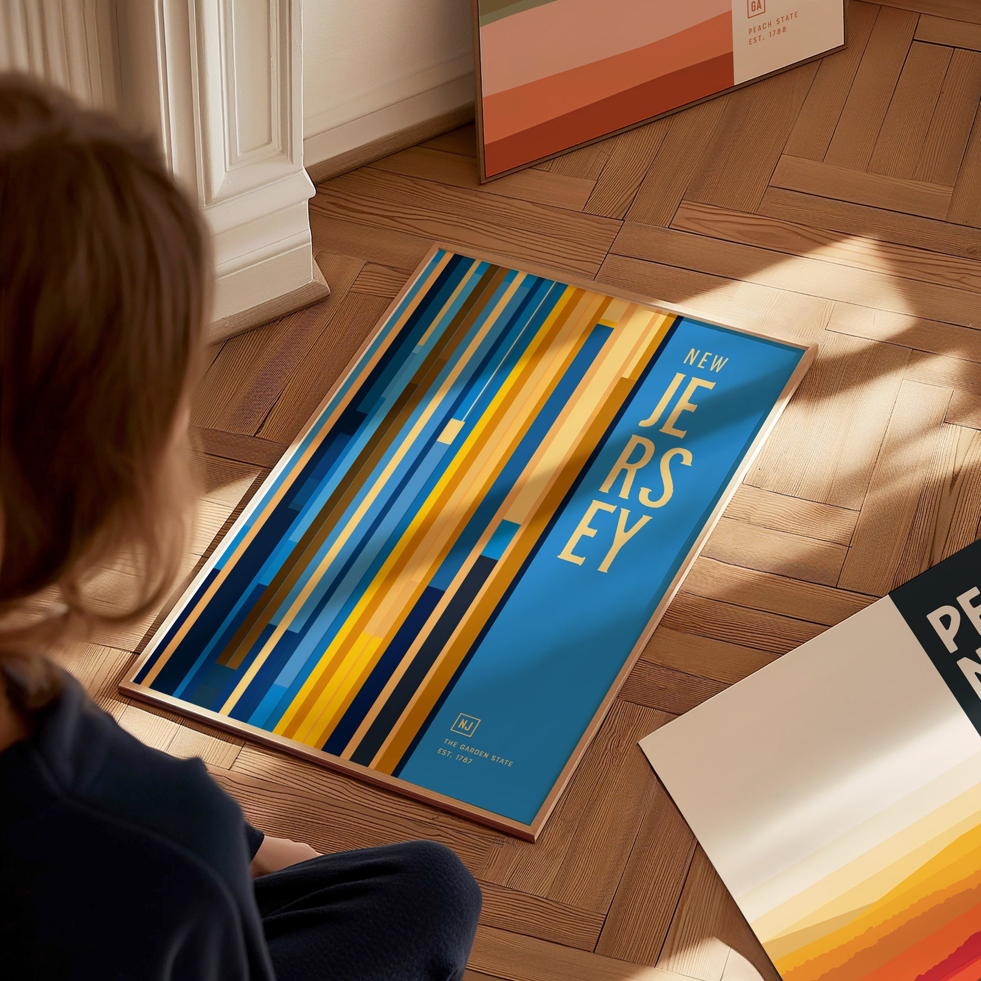 A person views a colorful New Jersey poster with vertical stripes in blue, yellow, and orange tones on a wooden floor. Other framed prints are partially visible nearby.
