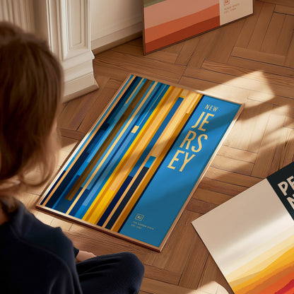 A person views a colorful New Jersey poster with vertical stripes in blue, yellow, and orange tones on a wooden floor. Other framed prints are partially visible nearby.