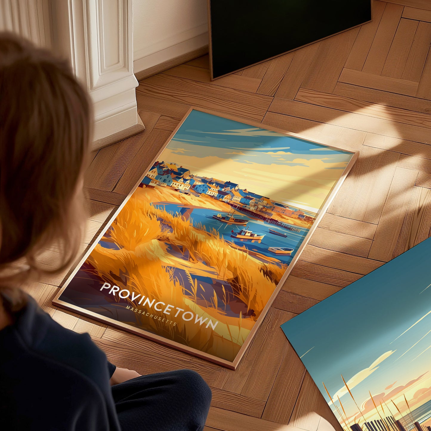 A child sits on a wooden floor, gazing at a framed travel print of Provincetown, depicting a vibrant harbor scene with colorful houses, boats, and golden grass under a bright sky. The artwork captures the lively essence of a seaside town.