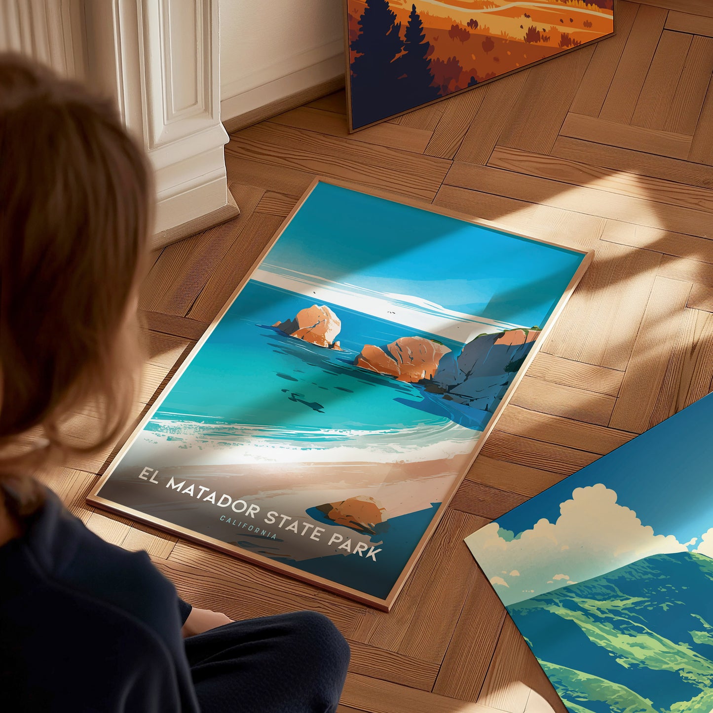 A person sits on a wooden floor, looking at a vibrant El Matador State Park poster featuring turquoise waters and rocky cliffs. Additional art pieces are visible nearby.