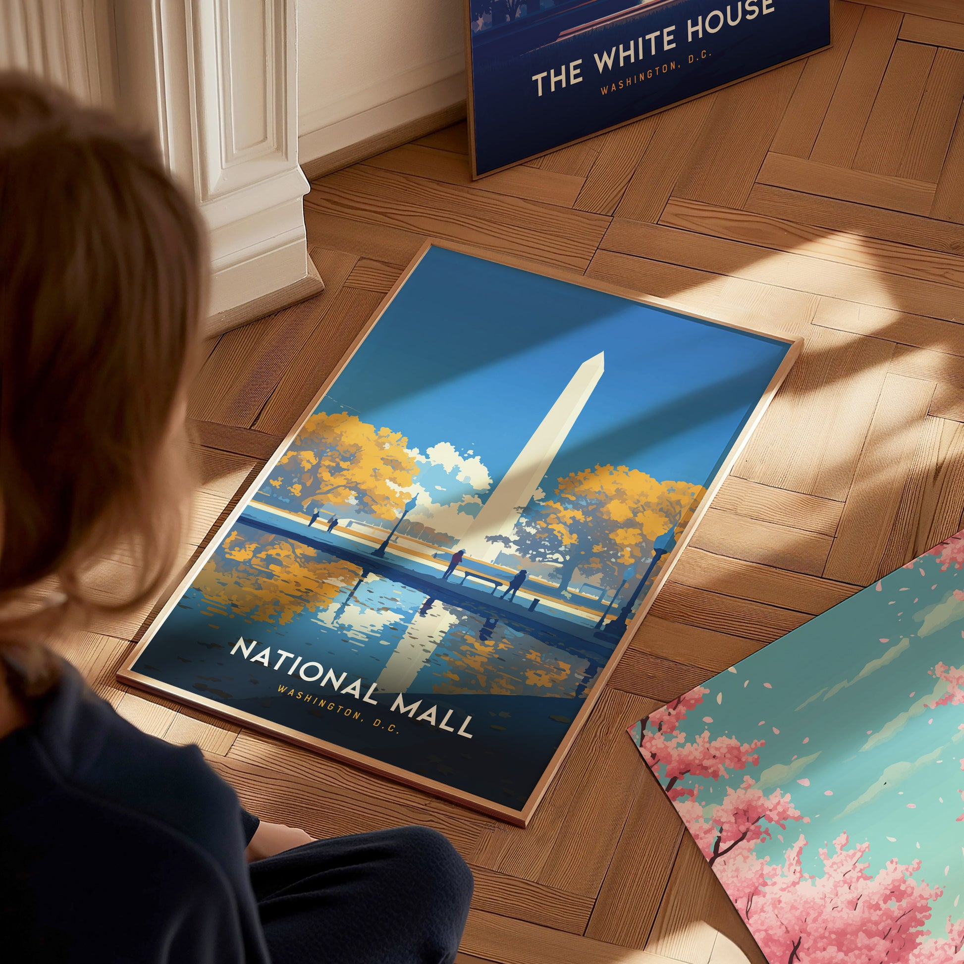 A person is looking at a framed poster of the National Mall with the Washington Monument, surrounded by autumn trees and a reflective pool, placed on a wooden floor.
