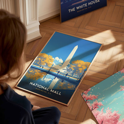 A person is looking at a framed poster of the National Mall with the Washington Monument, surrounded by autumn trees and a reflective pool, placed on a wooden floor.