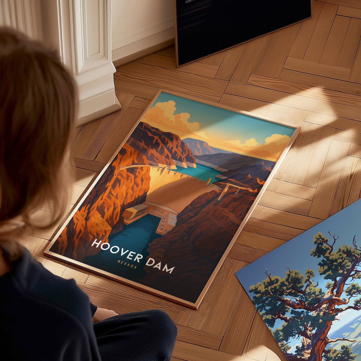 A person is sitting on a wooden floor looking at a framed poster of the Hoover Dam, depicted in vibrant desert tones with the Colorado River running through a canyon. Another colorful poster featuring a tree is partially visible beside it.