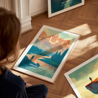 A person observes a framed Grand Teton National Park poster on a wooden floor, depicting a fisherman by a lake with mountains in the background.